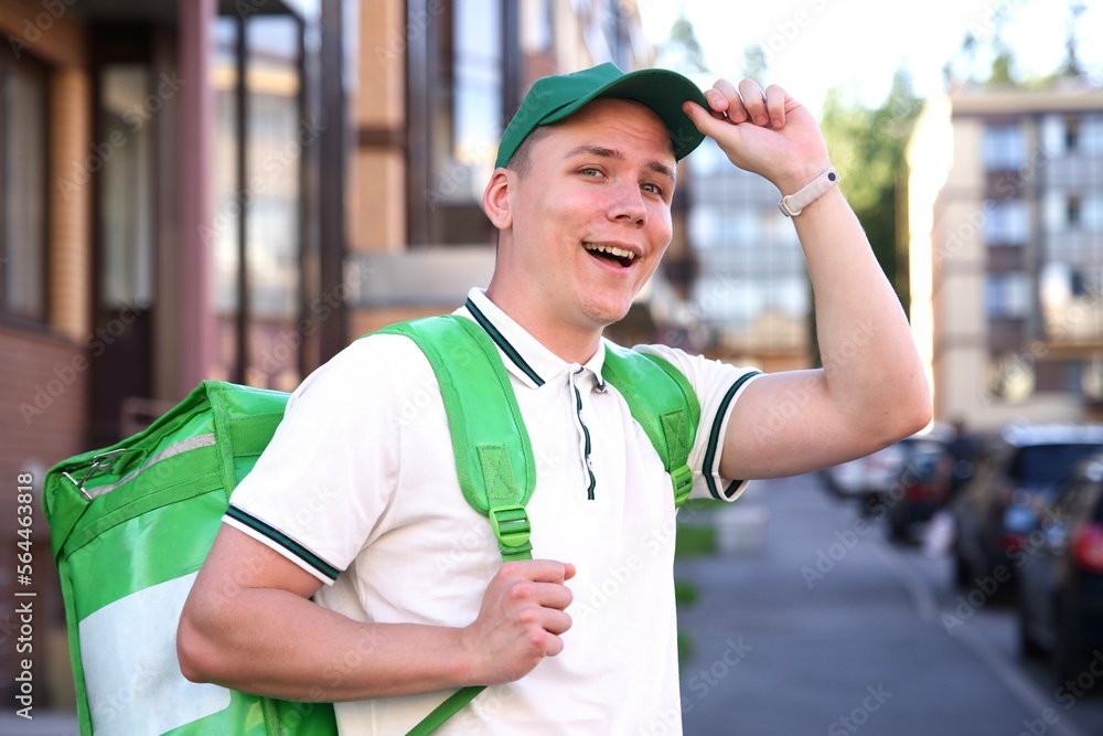 portrait of young handsome happy courier guy smile, food delivery man with green thermo box for food delivering food outdoors in the yard on buildings background at summer day in cap and uniform. 