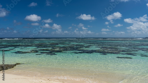 Corals are visible through the clear turquoise water of the ocean. The foam of waves over the reef in the distance.  White sand of the beach. Clouds in the blue sky. Seychelles. La Digue. 