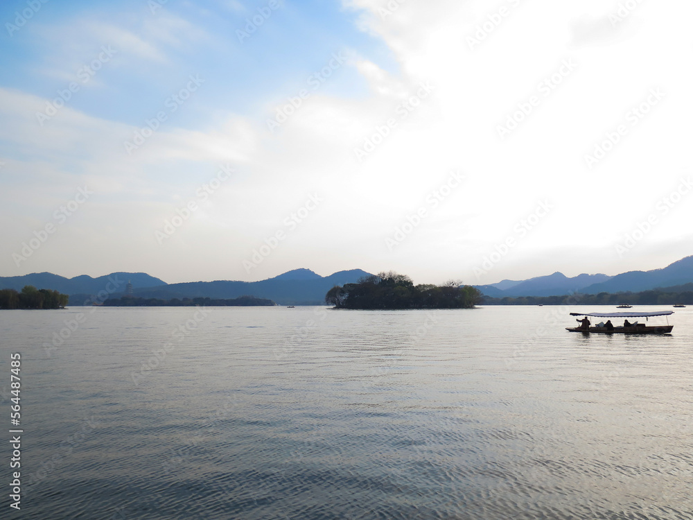A silhouette island and boat sailing on the calm sea at sunset in the background of mountains. Beautiful sky. For travel, background and blog etc. concepts. West Lake. Hangzhou city China A silhouette island and boat sailing on the calm sea at sunset in the background of mountains. Beautiful sky. For travel, background and blog etc. concepts. West Lake. Hangzhou city China
