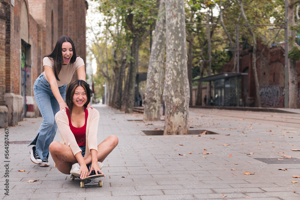 young woman laughing and having a good time pushing her friend sitting on a skateboard down a city street, concept of friendship and teenager lifestyle, copy space for text young woman laughing and having a good time pushing her friend sitting on a skateboard down a city street, concept of friendship and teenager lifestyle, copy space for text