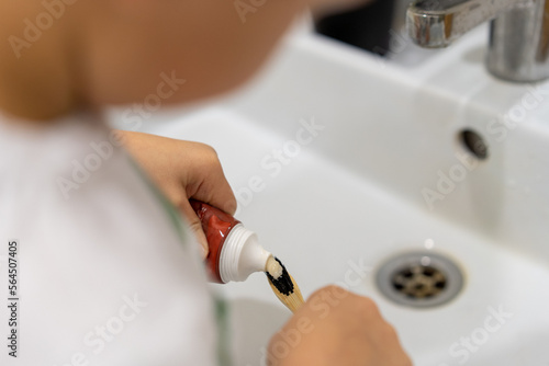 child squeeze tooth paste onto bamboo brush over white sink in bathroom