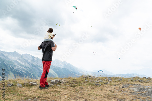 Rear view of a standing father with her little daughter on shoulders looking at a group of paragliders flying on a cloudy day at French alps .