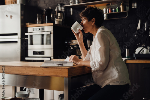 Senior woman drinking coffee and reading magazine while sitting in kitchen