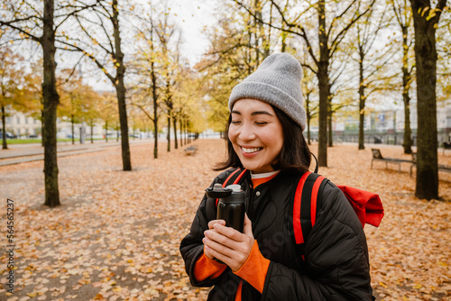 Smiling young asian woman drinking hot tea from thermos during walk in park
