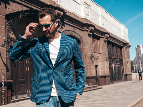 Handsome confident stylish hipster lambersexual model. Sexy modern man dressed in elegant blue suit and jeans. Fashion male posing in the street background in Europe city at sunset. In sunglasses