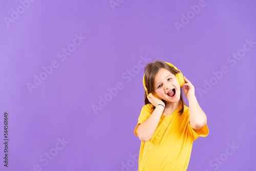 Smiling cute little girl listening to music on headphones and looking at camera isolated on purple background with copy space