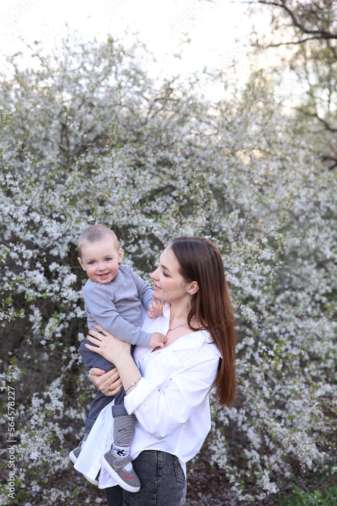 mother hugs her little son in spring near a blooming tree. Spring and plum blossom. Mother's Day. The love of a son and a mother. Gray clothes for a one-year-old boy. Kiss the son. child's laughter