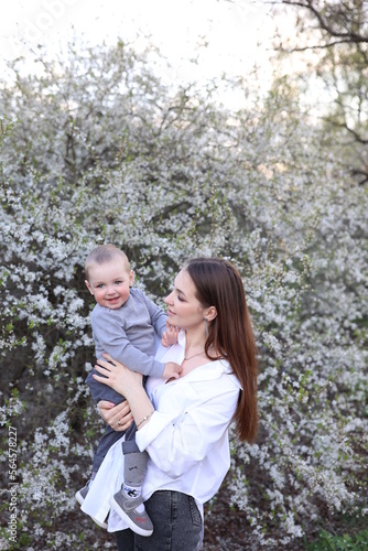 mother hugs her little son in spring near a blooming tree. Spring and plum blossom. Mother's Day. The love of a son and a mother. Gray clothes for a one-year-old boy. Kiss the son. child's laughter © Anhelina Tyshkovets