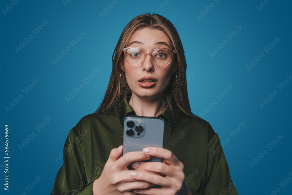Shocked woman reacting on sms frustrating on mobile phone, complaining on smartphone text, standing over blue background. Shocked woman reacting on sms frustrating on mobile phone, complaining on smartphone text, standing over blue background.