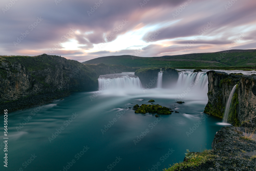 Fototapeta premium Long exposure sunset landscape over the icelandic Godafoss waterfall with blurred water falling into turquoise river and cloudy majestic sky