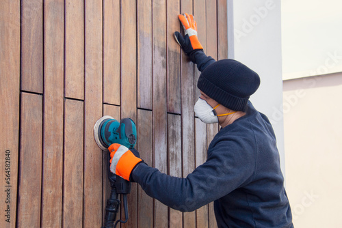 Man in dust mask sanding teak cladding on the house facade, exterior wood siding renovation photo