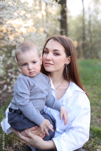 mother hugs her little son in spring near a blooming tree. Spring and plum blossom. Mother's Day. The love of a son and a mother. Gray clothes for a one-year-old boy. Kiss the son. child's laughter © Anhelina Tyshkovets