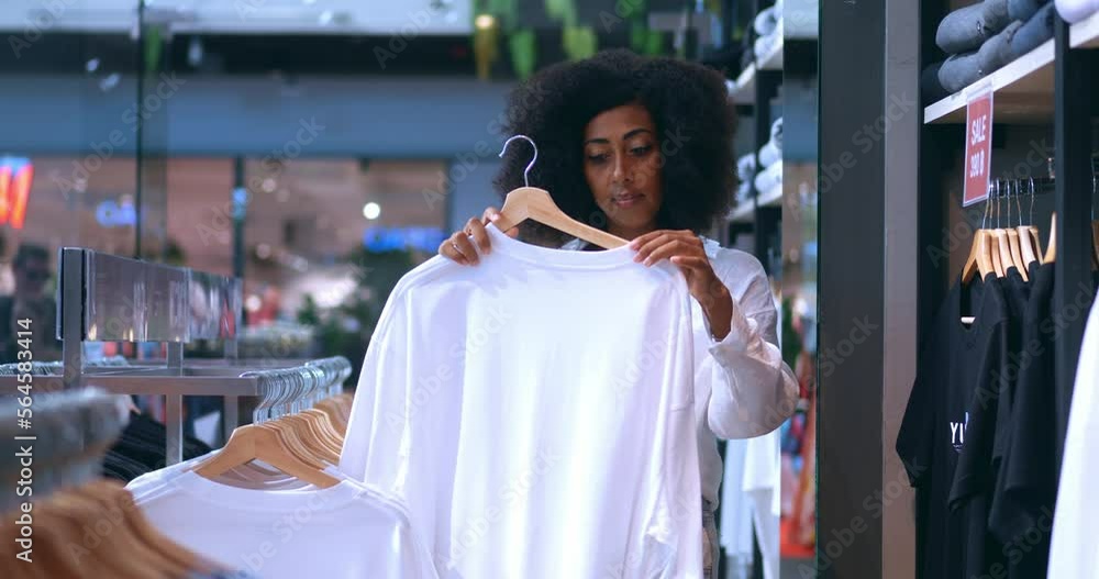 Happy young African American woman customer with shaggy hairstyle, in casual wear, holding stylish white t-shirt, smiling while enjoying shopping on her free time, in a modern shopping mall. Happy young African American woman customer with shaggy hairstyle, in casual wear, holding stylish white t-shirt, smiling while enjoying shopping on her free time, in a modern shopping mall.