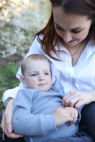 mother hugs her little son in spring near a blooming tree. Spring and plum blossom. Mother's Day. The love of a son and a mother. Gray clothes for a one-year-old boy. Kiss the son. child's laughter