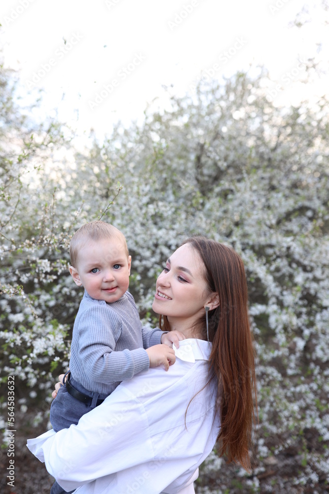 mother hugs her little son in spring near a blooming tree. Spring and plum blossom. Mother's Day. The love of a son and a mother. Gray clothes for a one-year-old boy. Kiss the son. child's laughter mother hugs her little son in spring near a blooming tree. Spring and plum blossom. Mother's Day. The love of a son and a mother. Gray clothes for a one-year-old boy. Kiss the son. child's laughter