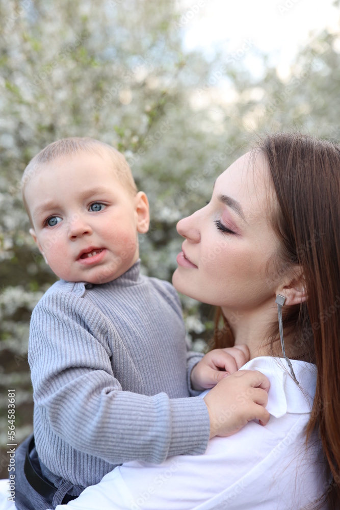 mother hugs her little son in spring near a blooming tree. Spring and plum blossom. Mother's Day. The love of a son and a mother. Gray clothes for a one-year-old boy. Kiss the son. child's laughter mother hugs her little son in spring near a blooming tree. Spring and plum blossom. Mother's Day. The love of a son and a mother. Gray clothes for a one-year-old boy. Kiss the son. child's laughter