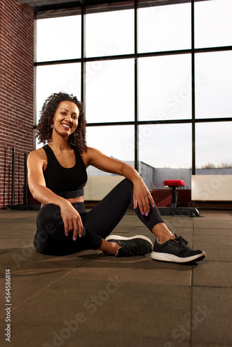 Attractive african american woman in sportswear looking at camera and smiling while sitting on the floor in a crossfit gym