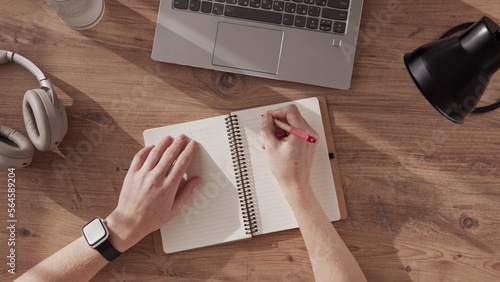 Top down view of a woman's hands writing in notebook and drawing. inscription in notebook. Work table with laptop, headphones and table lamp.