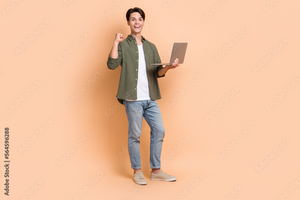 Full body size photo of young man work freelance it programmer professional first code done fist up hold netbook isolated on beige color background Full body size photo of young man work freelance it programmer professional first code done fist up hold netbook isolated on beige color background