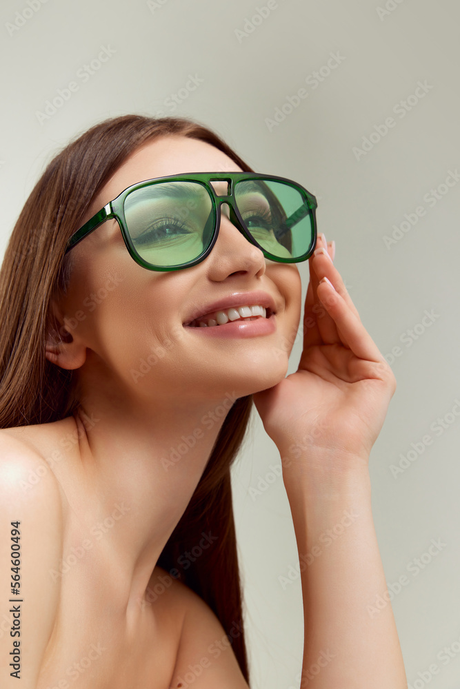 Portrait of young beautiful girl in green sunglasses posing, smiling over grey studio background. Perfect smile. Concept of natural beauty, youth, fashion, cosmetology, wellness, makeup, dental care Portrait of young beautiful girl in green sunglasses posing, smiling over grey studio background. Perfect smile. Concept of natural beauty, youth, fashion, cosmetology, wellness, makeup, dental care