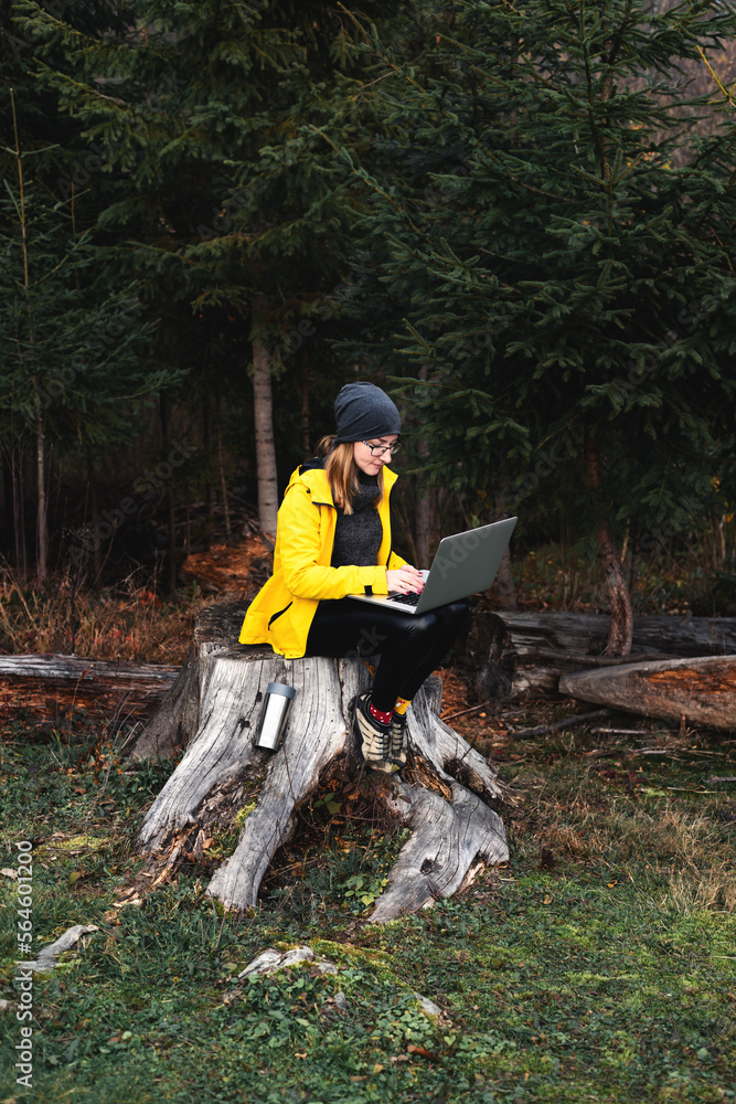 Woman wearing glasses and yellow jacket working on laptop outdoors surrounded by beautiful nature, forest and mountains. Workation concept. Freelancer work, remote work Woman wearing glasses and yellow jacket working on laptop outdoors surrounded by beautiful nature, forest and mountains. Workation concept. Freelancer work, remote work
