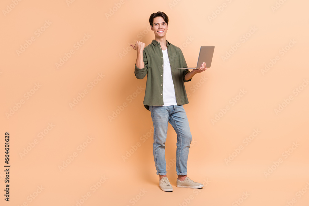 Full body length photo of young man interested looking empty space pointing finger with netbook promotion ad isolated on beige color background Full body length photo of young man interested looking empty space pointing finger with netbook promotion ad isolated on beige color background