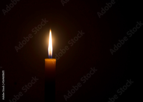 Single burning candle flame or light glowing on an orange candle on black or dark background on table in church for Christmas, funeral or memorial service with copy space.