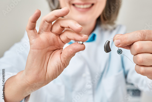 Hearing aid battery replacement. Positive audiologist showing hearing aid and battery for it, holding them in her hands in front of him photo