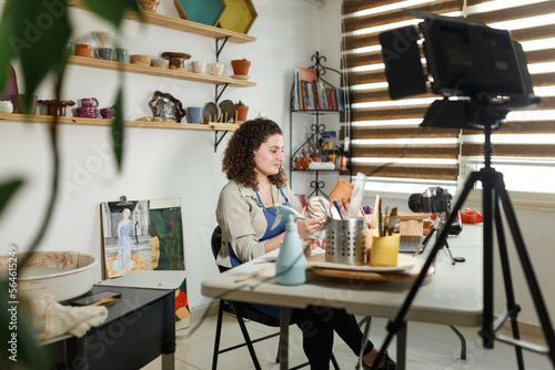Females potter in blue apron making a vlog, recording online course, clay master class,lessons in her studion with earthenware shelf on background.Using led lamp,studio equipment