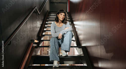 European pretty woman on the stairs sitting, looking at camera. Young brunette with short hair keep hand crossed in casual clothes. Indoor concept 