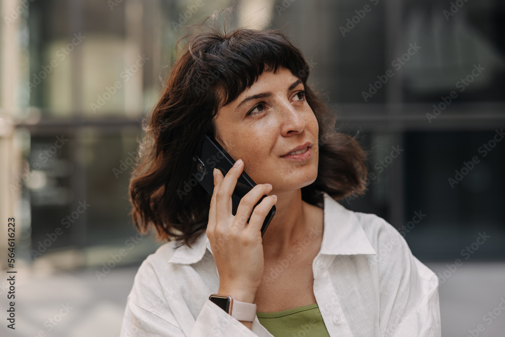 Image of caucasian pretty woman staying on the blur background with phone. Young brunette in hands cellphone and talking. Concept of use telephone technology Image of caucasian pretty woman staying on the blur background with phone. Young brunette in hands cellphone and talking. Concept of use telephone technology