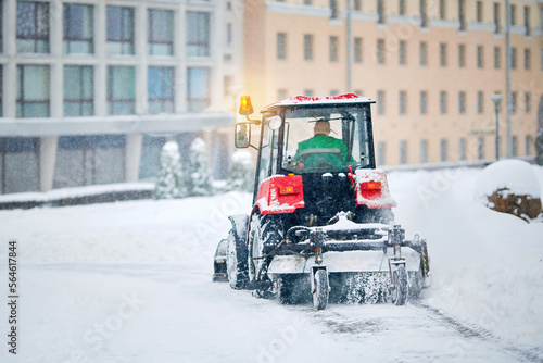 Tractor sweep snow, plow snow with rotating brush and snowplow from sidewalk. Snow plow vehicle clean snowy road, winter road maintenance during blizzard. Tractor clear snowy street