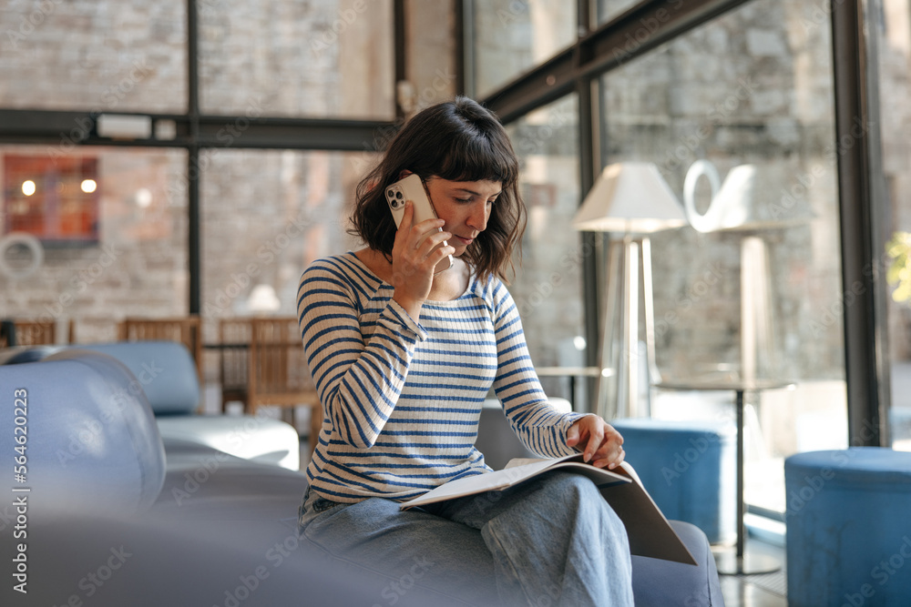 European business woman talking on cellphone and look at workbooks. Sitting in coworking place, wearing blue stripes shirt, jeans. Freelance work concept 