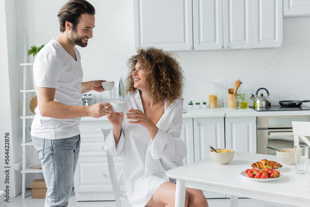 happy young man giving cup of coffee to curly girlfriend during breakfast in kitchen. happy young man giving cup of coffee to curly girlfriend during breakfast in kitchen.