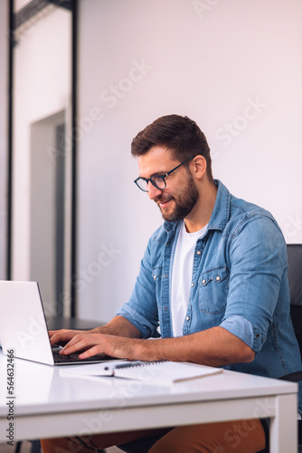 A smiling man with glasses is using a laptop in the office while sitting at his desk.