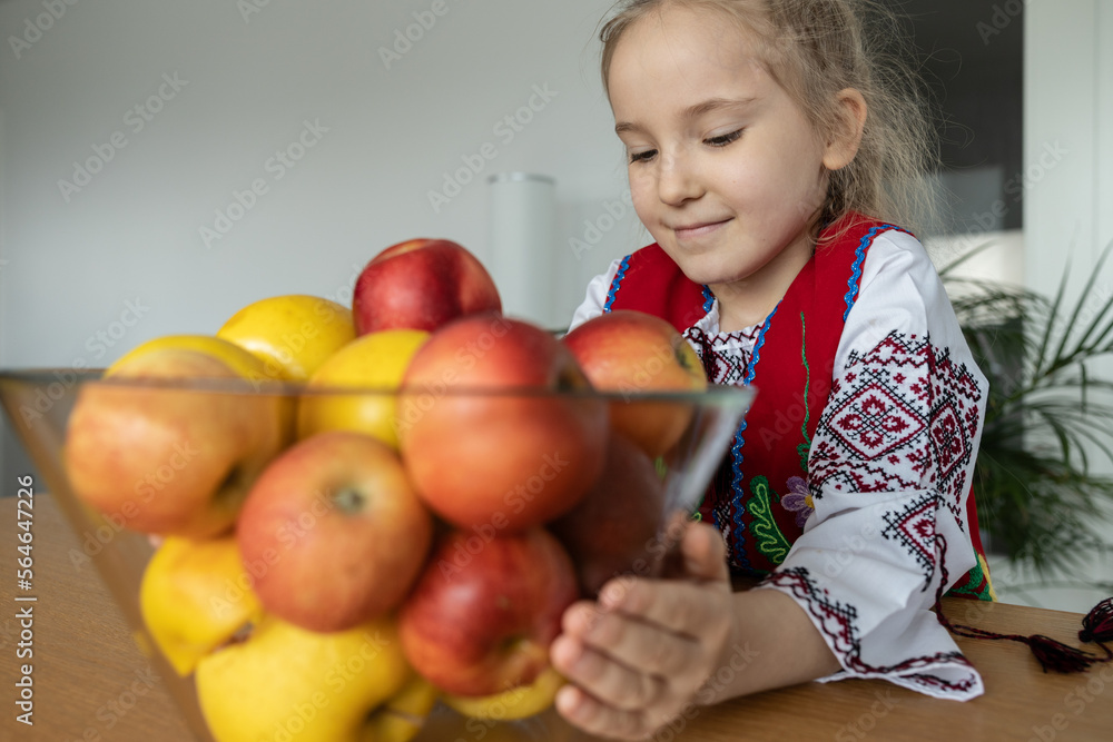 Fototapeta premium A little girl in a national Ukrainian costume stands at the table and holds a vase with bright apples in her hands.