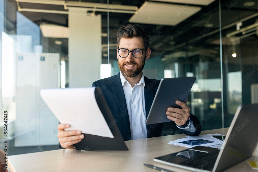 Cheerful middle aged businessman with documents and tablet working in office interior, free space Cheerful middle aged businessman with documents and tablet working in office interior, free space