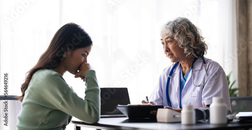 Woman senior doctor is Reading Medical History of Female Patient and Speaking with Her During Consultation in a Health Clinic. Physician in Lab Coat Sitting Behind a Laptop in Hospital Office.
