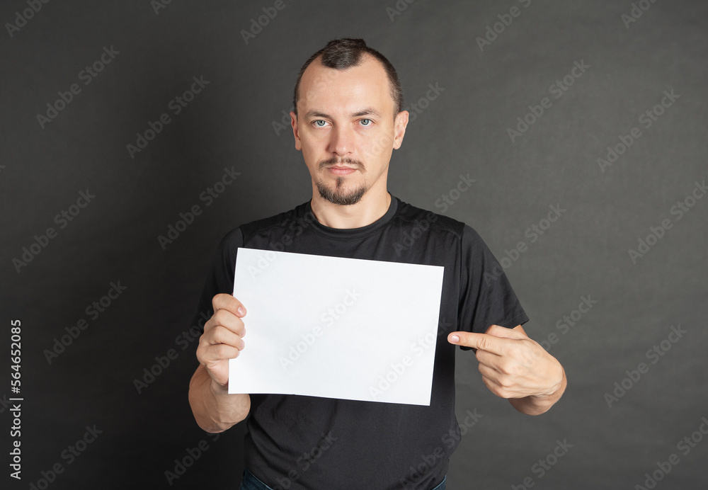 Young handsome man holding empty white sheet of paper and showing on it by his finger Young handsome man holding empty white sheet of paper and showing on it by his finger