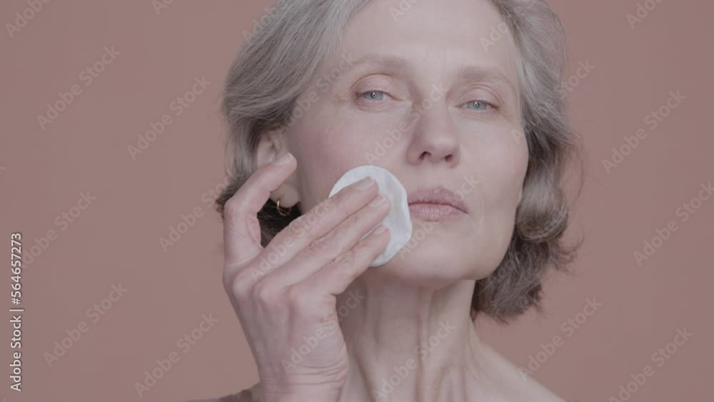 CU Portrait of very beautiful grey-haired 60s mature senior adult female doing her skincare routine. Using cotton pads. Studio shot, soft lighting. No make up, clean skin. Shot with ARRI Alexa Mini LF CU Portrait of very beautiful grey-haired 60s mature senior adult female doing her skincare routine. Using cotton pads. Studio shot, soft lighting. No make up, clean skin. Shot with ARRI Alexa Mini LF