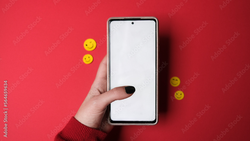 Mano femenina usando el móvil con pantalla blanca en fondo rojo con emoticonos Mano femenina usando el móvil con pantalla blanca en fondo rojo con emoticonos