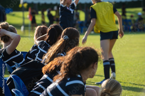 Substitute (bench, backups, interchange, reserves) girl players watching their team playing on a sideline waiting for their turn to play in an outdoor sport: soccer/football, rugby tournament.