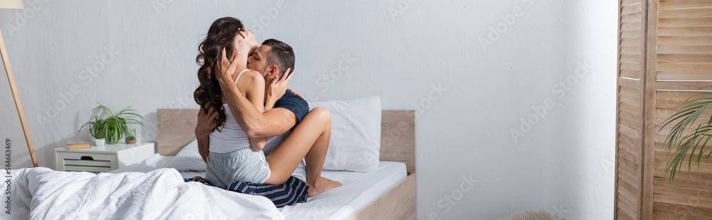 Young man in pajama kissing sexy girlfriend on bed at home, banner.