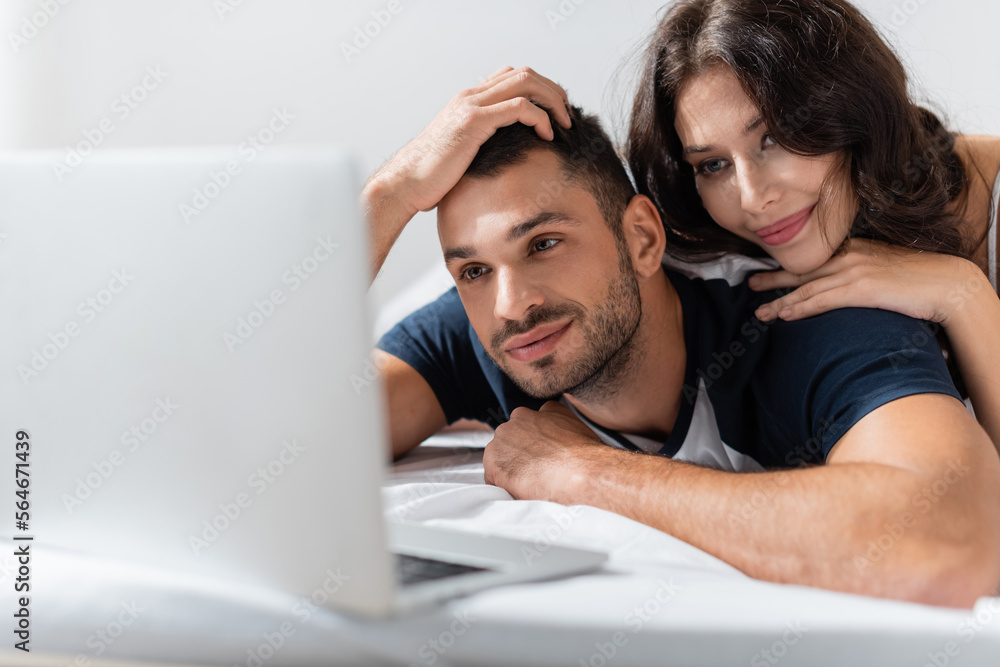 Smiling woman hugging boyfriend near blurred laptop on bed. Smiling woman hugging boyfriend near blurred laptop on bed.
