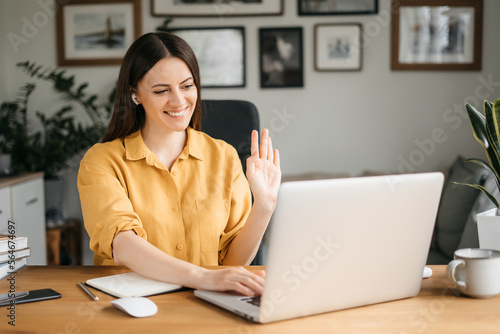 Happy young business woman employee, hr manager having remote online work hybrid meeting or distance job interview waving hand looking at laptop during virtual video conference call in office