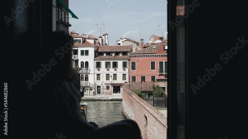 Caucasian girl sits on the windowsill and looking at the narrow canal in Venice. The girl enjoys a glass of wine on vacation. Canal view in Venice