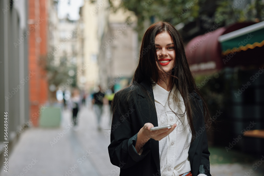 Business brunette woman with red lips smile with teeth with a phone in her hands, white shirt and black jacket fashion on the street, summer trip, vacation in the city tourist