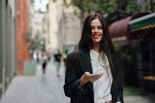 Business brunette woman with red lips smile with teeth with a phone in her hands, white shirt and black jacket fashion on the street, summer trip, vacation in the city tourist © SHOTPRIME STUDIO