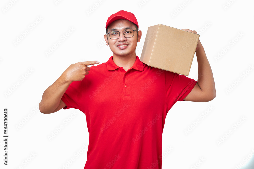 Friendly asian courier wearing red shirt and cap holding box on shoulder and pointing at package