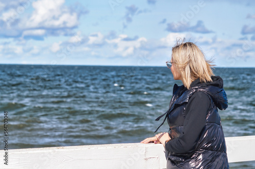 A blond-haired woman looks straight ahead into the endless sea. She has glasses and is wearing a black sweatshirt and a black warmer. She is standing with her hands on the railing of the pier. 